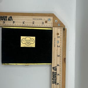 May include: A black playing card box with gold trim and a gold label that reads "CONGRESS Playing Cards Made in USA Or Spain." The box is next to a wooden ruler. The ruler measures in inches, equivalent to centimetres.
