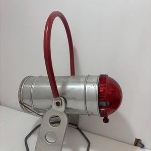 May include: Vintage silver-toned lantern with a red handle and a red lens. The lantern has a cylindrical body and a metal stand. The handle arches over the top. The lens is a rounded shape. The lantern is designed to be portable.