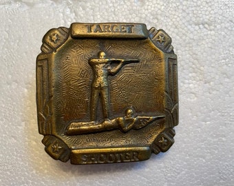 May include: Vintage brass-toned metal badge with the words "TARGET SHOOTER" and illustrations of two figures aiming rifles. The badge has a rectangular shape with decorative edges and star accents.