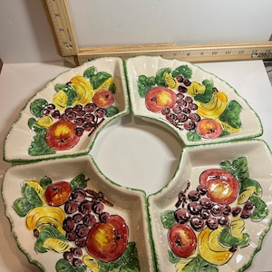 May include: A set of four off-white ceramic serving dishes arranged in a circular pattern. Each dish features a hand-painted design of red apples, purple grapes, and yellow bananas with green leaves. The edges are scalloped and trimmed in green.