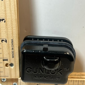 Puede incluir: Un objeto rectangular negro con la palabra "GUNLOK" y "PATENT NO. 5,437,119" impresas. El objeto mide aproximadamente 5 cm de largo y 4 cm de ancho. Está junto a una regla de madera.