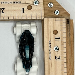 May include: A small, black and teal toy car with white wheels is positioned next to a wooden ruler. The ruler is marked with inch measurements and the text "MADE IN CHINA".