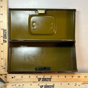 Pode incluir: Uma caixa de metal verde-oliva aberta com tampa articulada. A caixa tem formato retangular com cantos arredondados e uma trava. O interior é da mesma cor que o exterior. A caixa está colocada sobre uma superfície branca com uma régua de madeira.