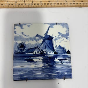 Peut inclure: Carreau de céramique carré avec une scène de moulin à vent hollandais bleu et blanc. L'œuvre représente un moulin à vent, des maisons et de l'eau, avec un code couleur bleu et blanc. Le carreau mesure environ 15 cm de côté.