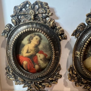 Puede incluir: Marco de fotos ovalado antiguo con un acabado de bronce detallado. El marco rodea una pequeña pintura de un querubín en tonos rojos y beige. El marco tiene detalles florales y de volutas ornamentados.