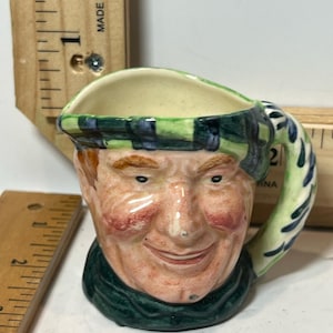May include: A small, ceramic mug shaped like a smiling man's head. The mug features a green and black plaid hat, a green scarf, and a handle with a blue and green floral design. The face is painted with rosy cheeks and red hair.