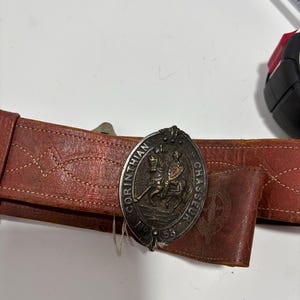 May include: A vintage brown leather belt with a decorative silver buckle. The buckle features an embossed design of two figures on horseback with the words "Corinthian Chasseur" and the number "53".