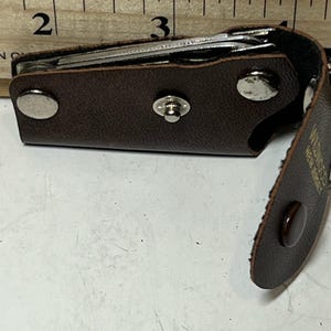 May include: A brown leather key holder with a snap closure, holding several keys. The holder is open, revealing the keys inside. The holder is next to a wooden ruler with inch measurements.
