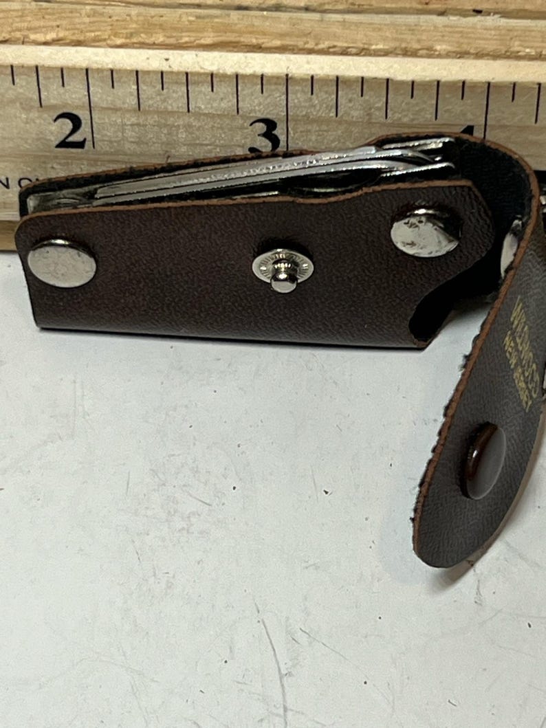 May include: A brown leather key holder with a snap closure, holding several keys. The holder is open, revealing the keys inside. The holder is next to a wooden ruler with inch measurements.