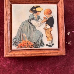May include: A framed tile featuring a vintage illustration of a woman in a blue dress and yellow hat giving a flower to a young boy in a black jacket and striped pants. A basket of flowers sits nearby. The frame is made of wood.