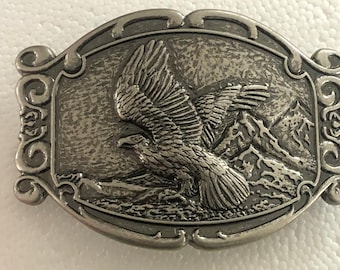 May include: A silver-colored metal belt buckle with an embossed design of an eagle in flight over a mountain range. The buckle has an ornate, scrolled border and a rectangular shape.