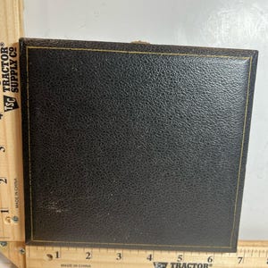 Könnte beinhalten: Eine schwarze, quadratische Box mit strukturierter Oberfläche und goldenem Rand. Die Box ist geschlossen und scheint aus Leder oder einem ähnlichen Material zu bestehen. Die Box liegt auf einem Holzlineal.