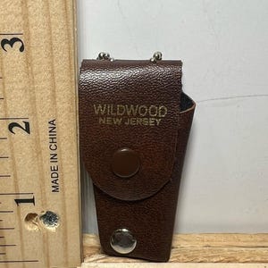 May include: A brown leatherette case with the text "WILDWOOD NEW JERSEY" in gold lettering. The case has a snap closure and a small loop at the top. The case is approximately 2 inches tall.