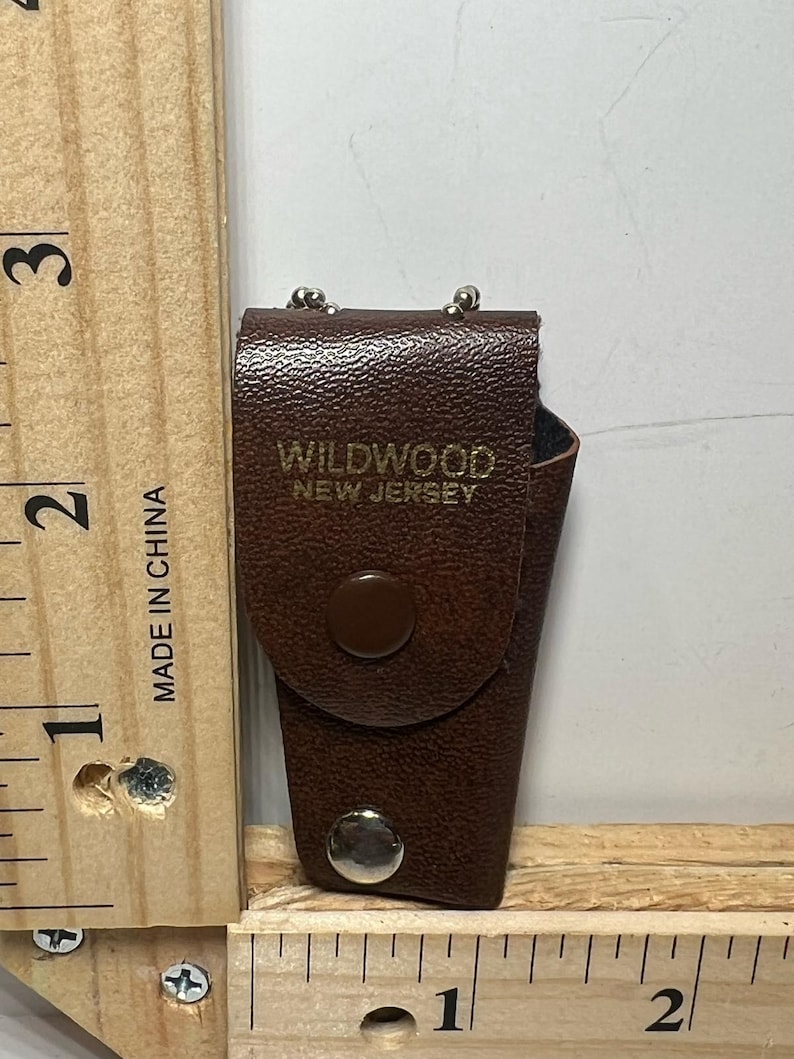May include: A brown leatherette case with the text "WILDWOOD NEW JERSEY" in gold lettering. The case has a snap closure and a small loop at the top. The case is approximately 2 inches tall.
