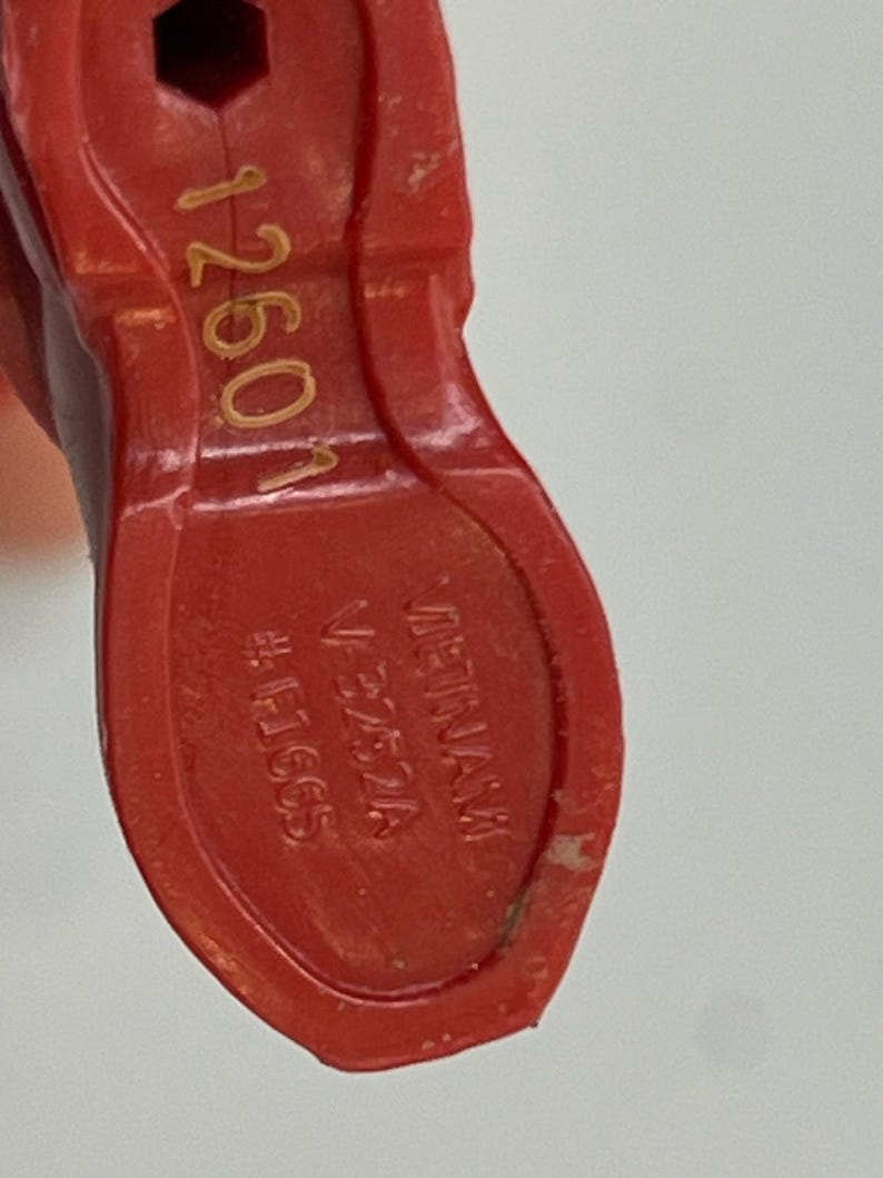 Puede incluir: Primer plano de un objeto de pl&aacute;stico rojo con el texto "1260N" en dorado. Tambi&eacute;n es visible el texto "VIETNAM" y "V-3752A". El objeto tiene forma de zapato y un agujero hexagonal.