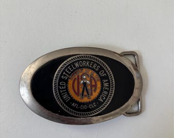 May include: Oval metal belt buckle with a black center featuring the text "UNITED STEELWORKERS OF AMERICA" around a circular emblem. The emblem includes a figure and the letters "USA".