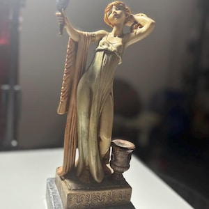 May include: A figurine of a person with reddish hair wearing a flowing, light-colored gown, holding a small hand mirror. The figure stands on a rectangular base with decorative carvings, positioned atop a dark, polished marble-like pedestal.