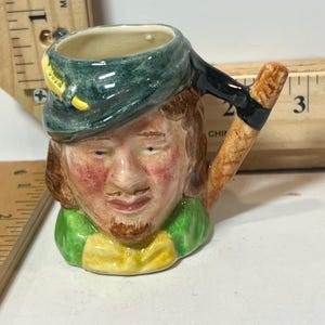 以下が含まれることがあります： 緑色の帽子と赤みがかった顔をした男性の頭の形をした小さなセラミックマグ。ハンドルは木製の斧のようにデザインされています。マグカップは鮮やかな色で塗装されています。