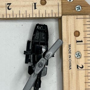 May include: A black toy helicopter with a gray propeller, positioned on a wooden ruler. The ruler is marked with inches and the text "MADE IN CHINA". The helicopter is approximately 2 inches long.