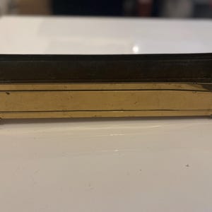 May include: A rectangular brass planter with decorative feet. The planter has a long, narrow shape with a dark interior and a polished exterior. The brass has a warm, golden hue, and the feet add a touch of elegance.