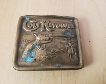 Könnte beinhalten: Vintage Gürtelschnalle aus Messing mit einem Colt-Revolver-Design. Die Schnalle trägt die geprägten Worte "COLT REVOLVER" sowie ein Bild eines Revolvers und eines Cowboys auf einem Pferd. Die Schnalle weist eine blau-grüne Patina auf.