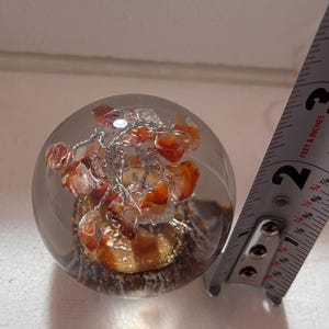 Puede incluir: Un pisapapeles de cristal transparente con un interior decorativo. El interior presenta un grupo de piedras naranjas y rojas y alambre plateado. El pisapapeles tiene aproximadamente 6,35 cm de diámetro.