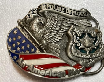 Puede incluir: Hebilla de cinturón plateada con imágenes patrióticas. Presenta un águila, la bandera estadounidense y una placa de policía. El texto dice "THE POLICE OFFICER" y "An American Hero". La hebilla es un artículo conmemorativo.