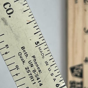 May include: Close-up of a vintage wooden ruler with black markings and text. The ruler is marked in inches and fractions of an inch, with the text "MADE IN U.S.A." and company information visible.