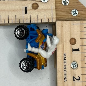 May include: A miniature toy motorcycle with a rider. The motorcycle is blue and gold, with black wheels. The rider is wearing a white suit with red accents. The toy is approximately 1 inch long.