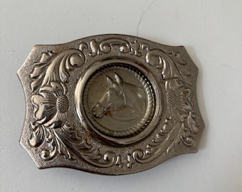 May include: Silver-toned metal belt buckle with a horse head design. The rectangular buckle features a central circular medallion with a horse profile, surrounded by a rope-like border. Floral and scrollwork details adorn the edges.