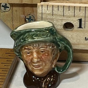 May include: A miniature ceramic jug shaped like a man's head, featuring a green hat and brown attire. The face is detailed with painted features. A wooden ruler is visible in the background. The jug has a handle.