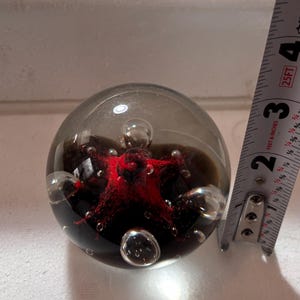 Puede incluir: Un pisapapeles de cristal transparente con un diseño rojo oscuro en forma de estrella en su interior. El pisapapeles es esférico y tiene varias burbujas de aire. Mide aproximadamente 7.6 cm de diámetro.
