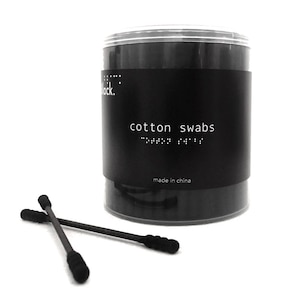 May include: A black plastic container of black cotton swabs. The container has a label that reads "cotton swabs" in white text. The container is sitting on a white surface. Two black cotton swabs are lying on the surface next to the container.
