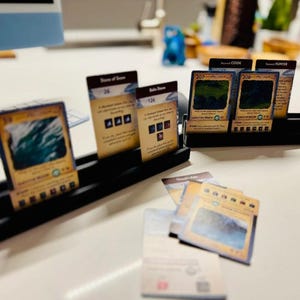 May include: A collection of game cards with various illustrations and text, displayed in black card holders. The cards feature images of landscapes and symbols, with text indicating game information. Some cards are face up, while others are in holders.