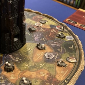 May include: A close-up of a board game with a brown, circular board and a black tower in the centre. The board is divided into hexagonal sections with different names, such as "The Cloister", "Howling Desert", and "Bone Hills". There are small white figures on the board.