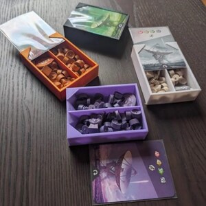May include: A collection of board game storage containers in various colors. The containers hold game pieces and cards. The containers are orange, black, white, and purple. The game pieces are brown, beige, and gray. The cards have images of a satellite dish and a flying object.
