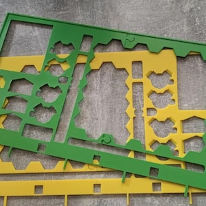 K&ouml;nnte beinhalten: Zwei Kunststoffschablonen, eine gr&uuml;ne und eine gelbe, liegen auf einer grauen Oberfl&auml;che. Die Schablonen haben verschiedene ausgeschnittene Formen, darunter geometrische Formen und vogel&auml;hnliche Designs. Die gr&uuml;ne Schablone liegt oben.