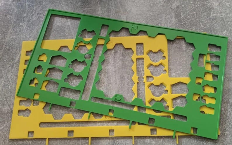 K&ouml;nnte beinhalten: Zwei Kunststoffschablonen, eine gr&uuml;ne und eine gelbe, liegen auf einer grauen Oberfl&auml;che. Die Schablonen haben verschiedene ausgeschnittene Formen, darunter geometrische Formen und vogel&auml;hnliche Designs. Die gr&uuml;ne Schablone liegt oben.