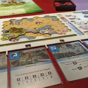 K&ouml;nnte beinhalten: Ein Brettspiel mit einem beigefarbenen Spielbrett, Karten und roten Spielsteinen. Das Spielbrett hat ein sechseckiges Design mit einer Landschaft. Die Karten haben Bilder und Text. Das Spiel befindet sich auf einer roten Oberfl&auml;che.