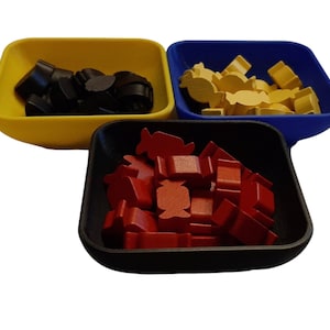 May include: Three square-shaped, colorful trays filled with game pieces. The trays are yellow, blue, and black. The yellow tray holds black pieces, the blue tray holds yellow pieces, and the black tray holds red pieces. The pieces are various shapes.