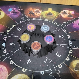 Può includere: Un'impostazione di gioco da tavolo con un tappetino nero a tema cosmico e la scritta "ARCS". Un supporto di plastica nero contiene pile di gettoni colorati. I gettoni hanno vari simboli e colori, tra cui viola, blu, giallo e arancione.