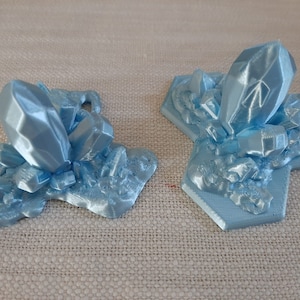 May include: Two 3D printed blue crystal formations on a light brown fabric background. The crystals are on hexagonal bases.