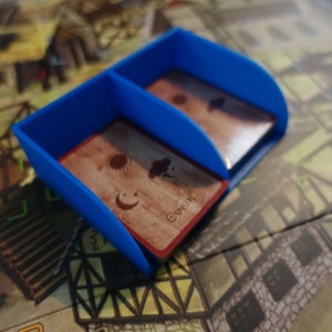 Pu&ograve; includere: Un porta carte in plastica blu con due scomparti. Il porta carte &egrave; appoggiato su un tabellone da gioco con una carta marrone con un'immagine di sole, luna e nuvola al suo interno. La carta ha anche la parola "Evento" stampata su di essa.