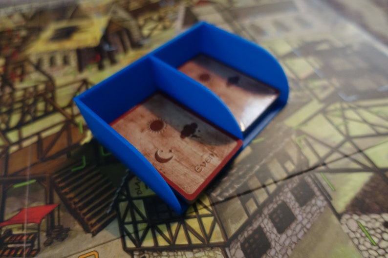 Pu&ograve; includere: Un porta carte in plastica blu con due scomparti. Il porta carte &egrave; appoggiato su un tabellone da gioco con una carta marrone con un'immagine di sole, luna e nuvola al suo interno. La carta ha anche la parola "Evento" stampata su di essa.