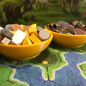 May include: Two yellow bowls, one filled with small wooden game pieces in various colors and shapes, the other filled with copper-colored coins. The bowls are sitting on a green and blue game board.