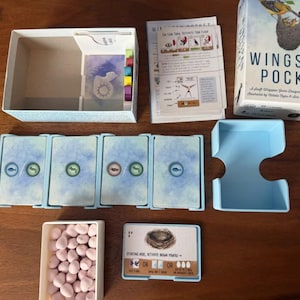 May include: Wingspan Pocket board game set including a white box, a light blue card holder, four stacks of blue-backed cards with circular icons, a small white tray filled with light pink egg tokens, a rule sheet, and a game board card.