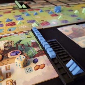 May include: A close-up of a board game with blue and green game pieces, dice, and a score tracker. The score tracker is a black plastic tray with blue plastic cubes. The game board features a colorful illustration of a wagon and a crow.