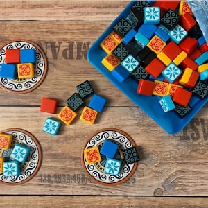 Può includere: Un gioco da tavolo con piastrelle quadrate colorate e sottobicchieri. Le piastrelle sono rosse, blu, gialle e nere con motivi decorativi. Un vassoio blu e un sacchetto a fantasia contengono le piastrelle. I sottobicchieri hanno un design simile.