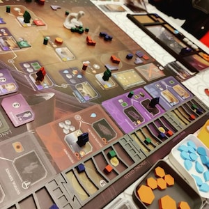 Puede incluir: Un juego de mesa en progreso, con un tablero detallado con cartas, piezas de juego de madera de varios colores y bandejas. El tablero tiene texto e ilustraciones, con un enfoque en la estrategia y la gestión de recursos.