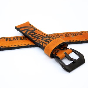Puede incluir: Una correa de reloj de cuero marrón con costuras negras y el texto "Rawlings ED™ SERIES PLAYER" impreso en ella. La correa tiene una hebilla de metal negro.